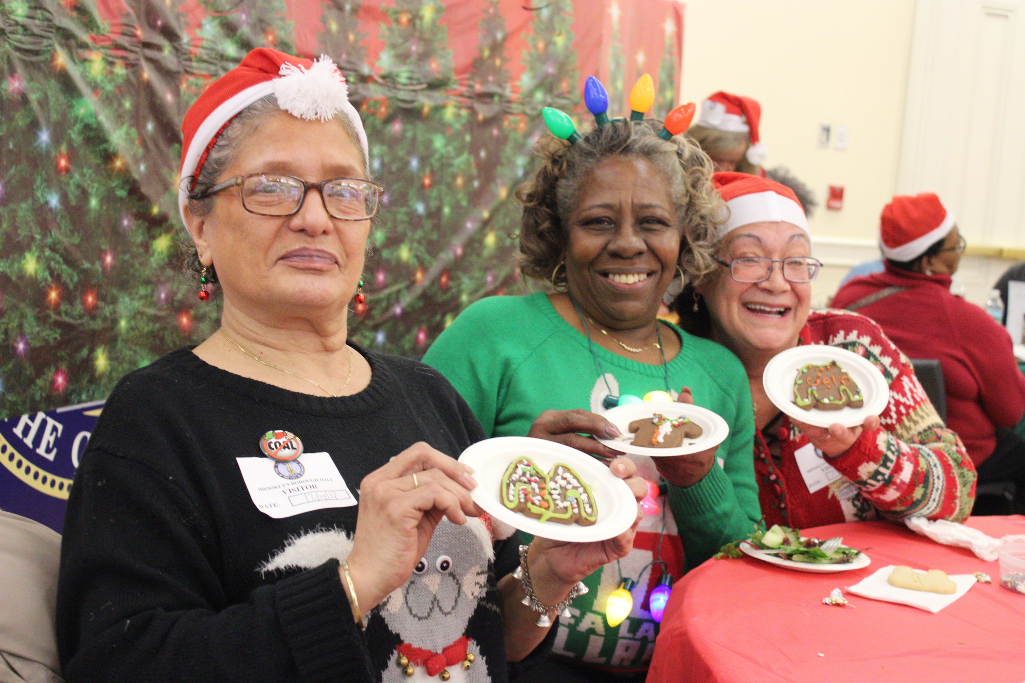 Three senior women in festive attire hold up holiday cookies and smile for camera