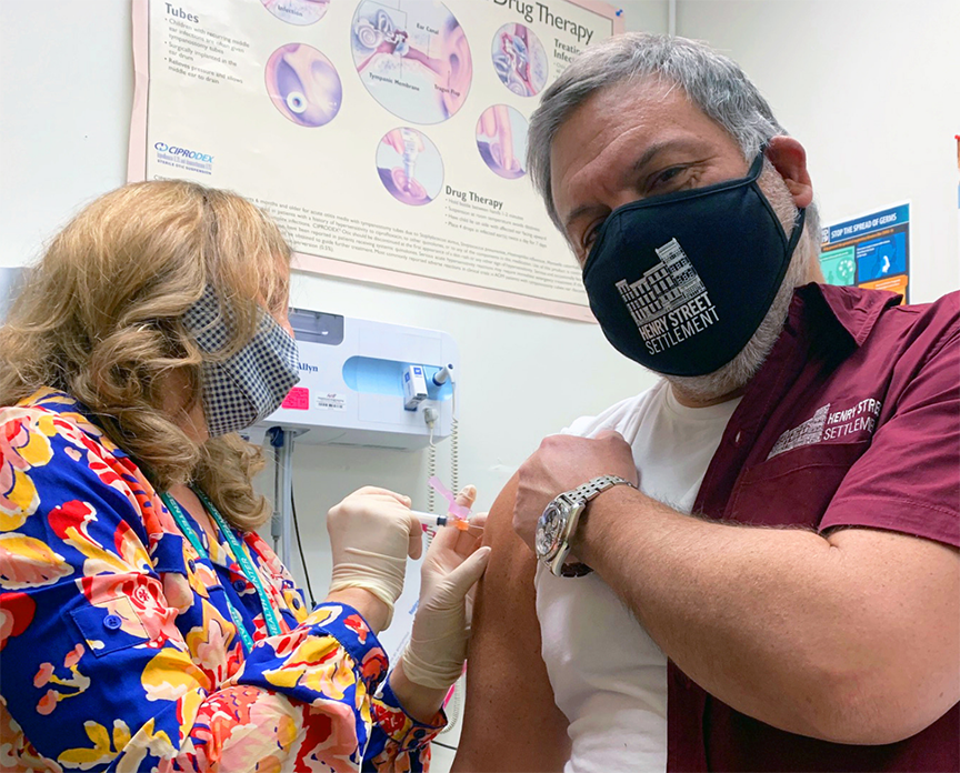 A woman wearing a mask and a blue, yellow, and red patterned shirt gives a COVID-19 vaccination to Henry Street Settlement President & CEO David Garza, who wears a maroon Henry Street Settlement shirt and black Henry Street Settlement mask