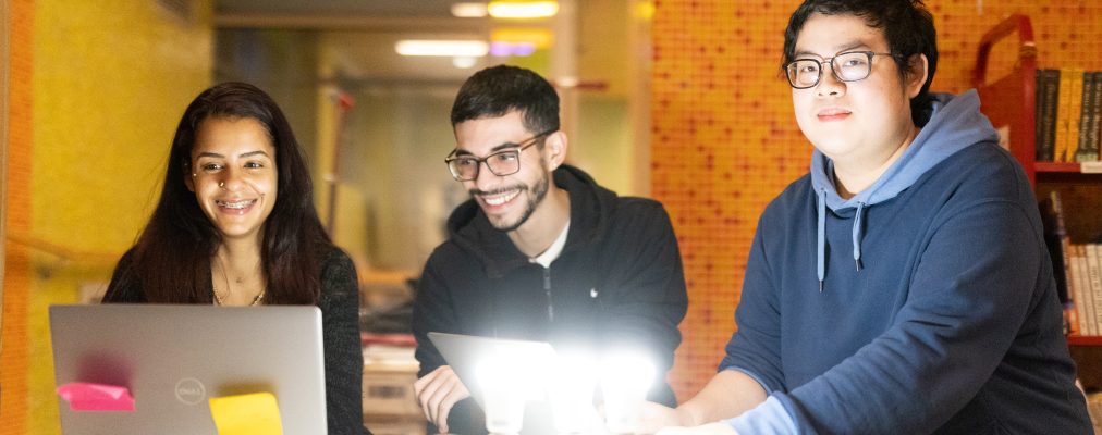 Three young adults collaborate in a warmly lit space. A smiling woman works on a laptop, while a man in a hoodie holds another. A person in a blue hoodie interacts with glowing light bulbs.